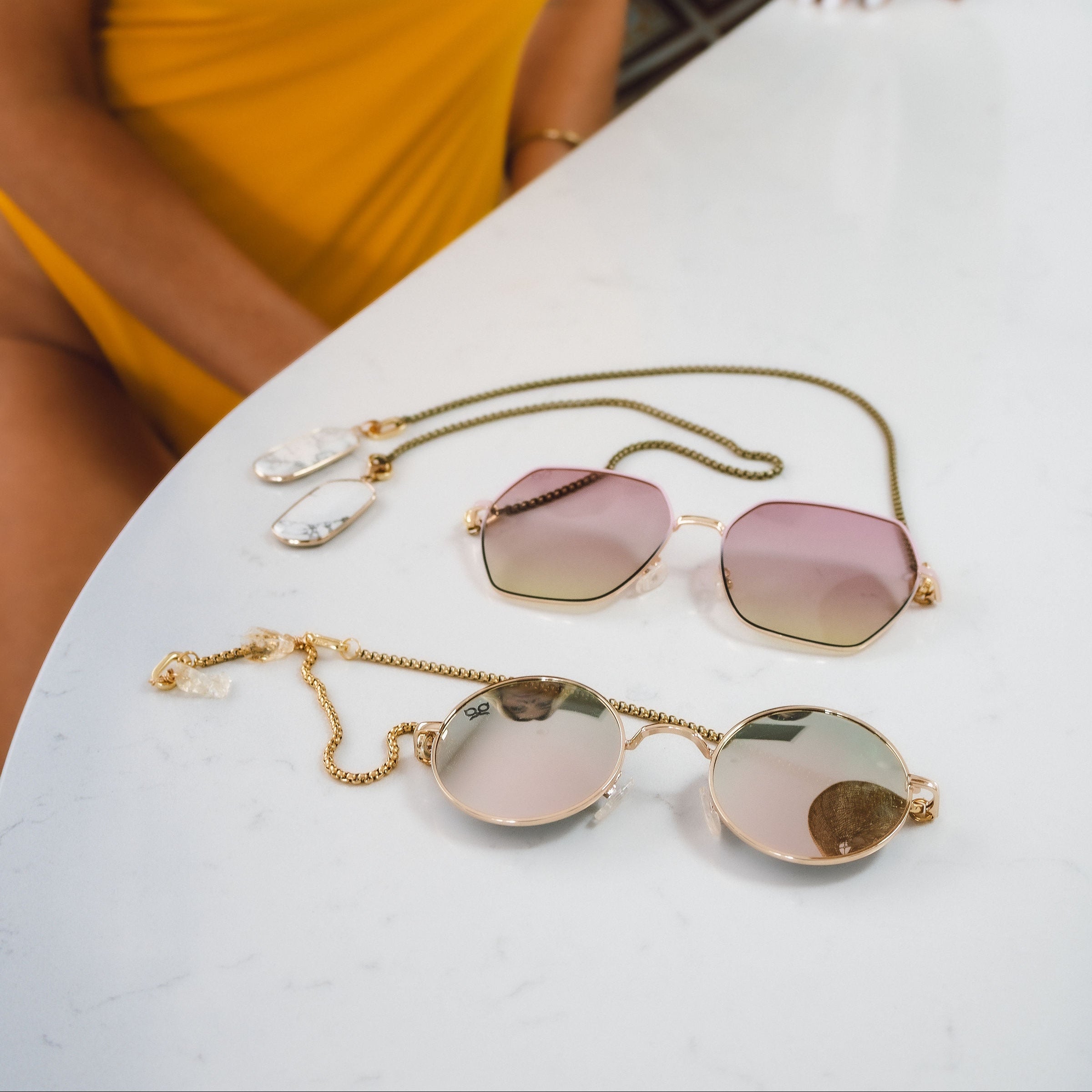 Two pairs of sunglasses on a white surface with a person in a yellow swimsuit partially visible.