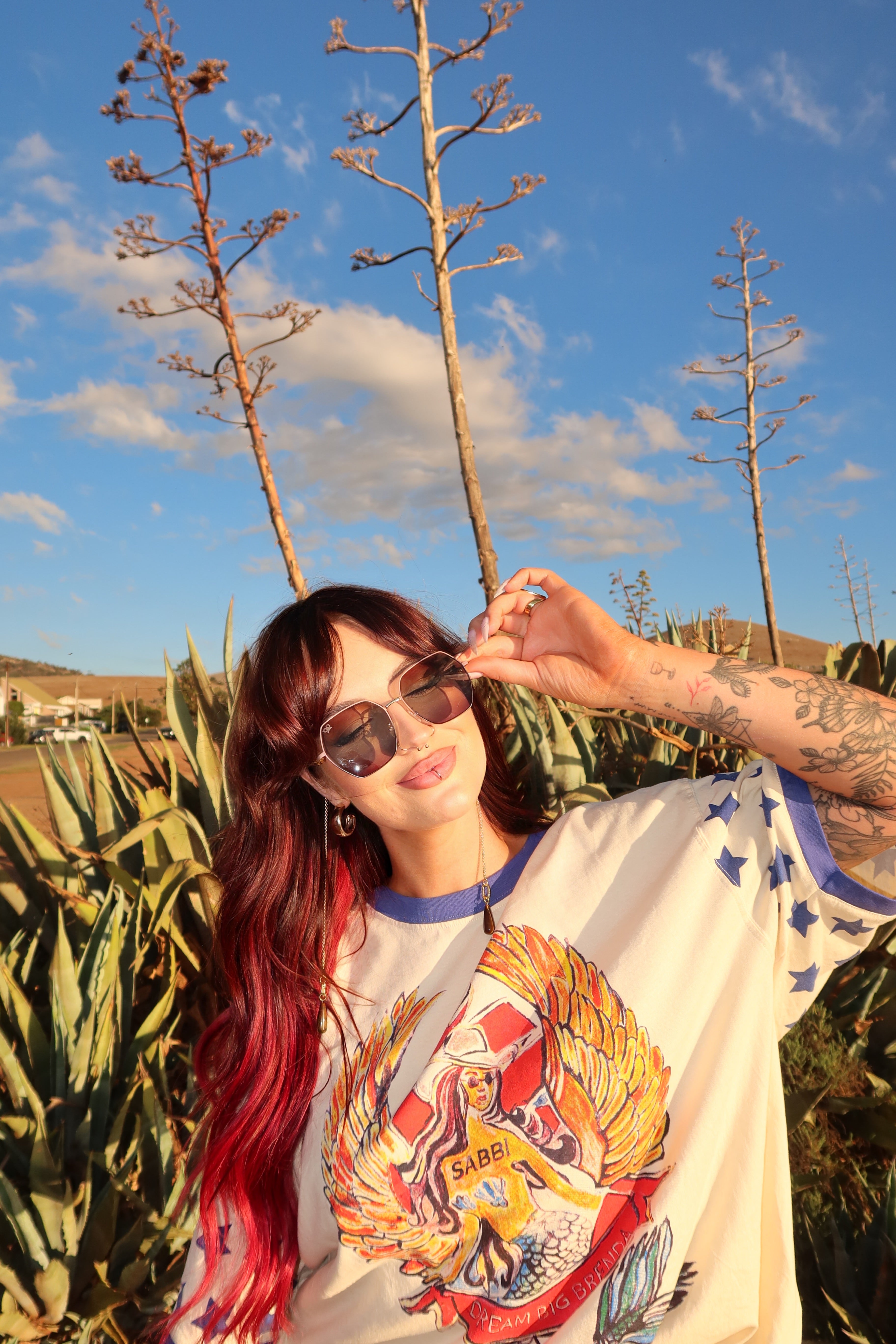 Person with red hair and tattoos wearing sunglasses and a colorful shirt in a natural setting.