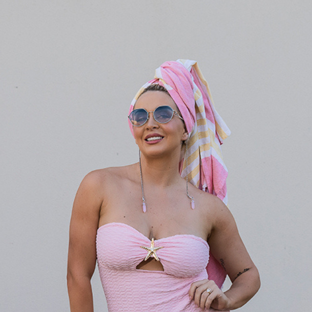 Woman in a pink dress and headscarf standing against a white wall with palm trees in the background. Wearing Ziggy Sunglasses