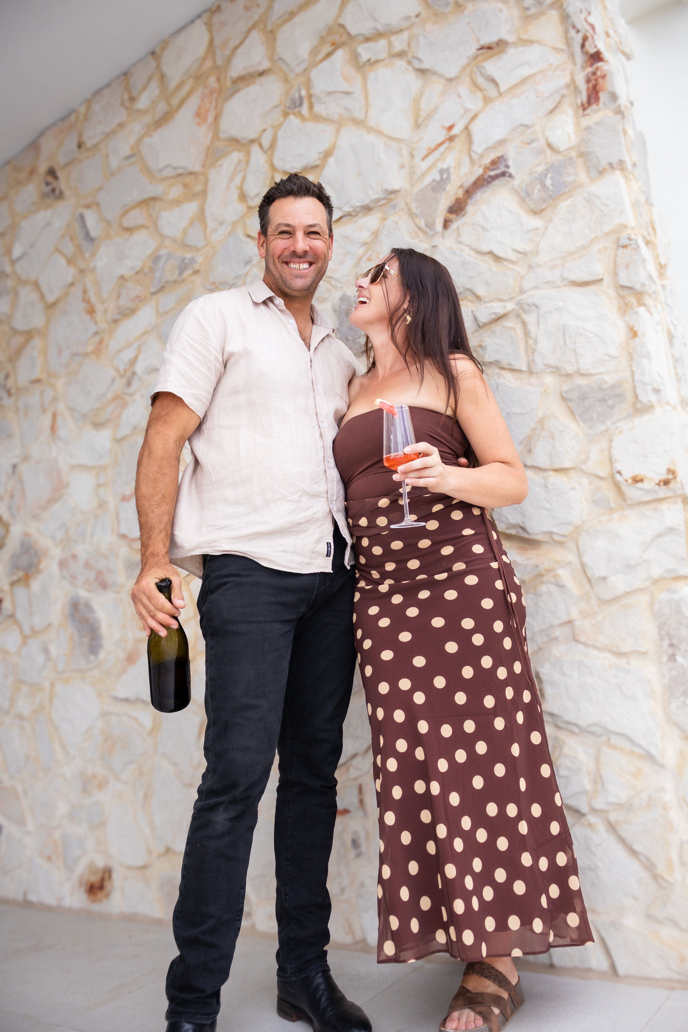 Man and woman standing together against a stone wall, holding drinks.